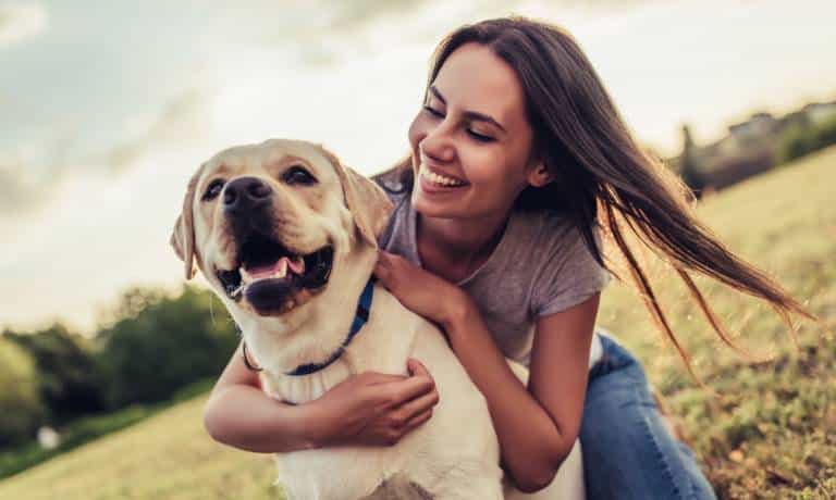 Unexpected Things You Need When Owning a Pet 1 A young woman in a purple shirt and jeans hugs her dog in a park. She and the dog are smiling and happy.