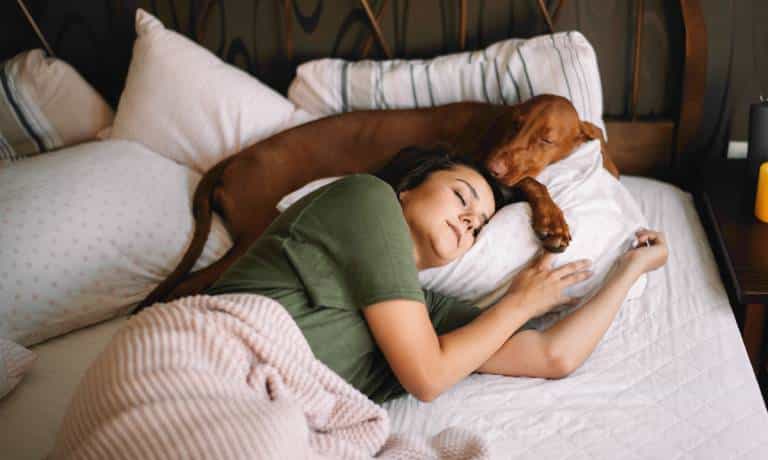 How To Stop Your Bedroom From Smelling Like Your Pets 1 A woman in a green shirt and a brown dog sleeping in the same bed and using the same white pillow.