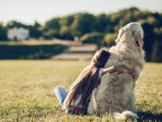 A child in a white shirt hugs a fluffy golden dog while sitting on the grass in an open field with trees in the background.