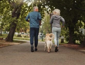 A mature couple jogging in a well-shaded park alongside their golden retriever, who is trotting between them.