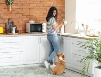 A young woman wearing a striped T-shirt and jeans drying a cup with a towel while looking down at a corgi next to her.