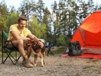 A man sits in a camping chair and pets a dog next to an orange tent set up in a large forest clearing.