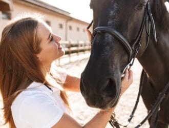 A smiling young woman stands outside and holds onto her horse's bridle as she looks up at the horse.