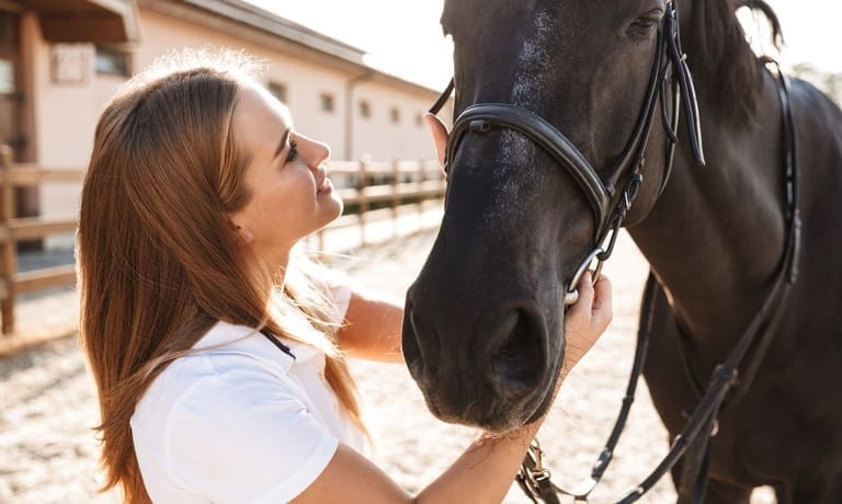 5 Essential Care Tips All Horse Owners Should Know 1 A smiling young woman stands outside and holds onto her horse's bridle as she looks up at the horse.