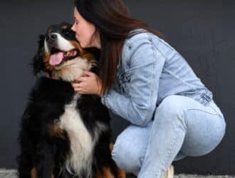 Woman in denim outfit kneeling and kissing a large black and brown dog with tongue out, sitting by a dark wall.