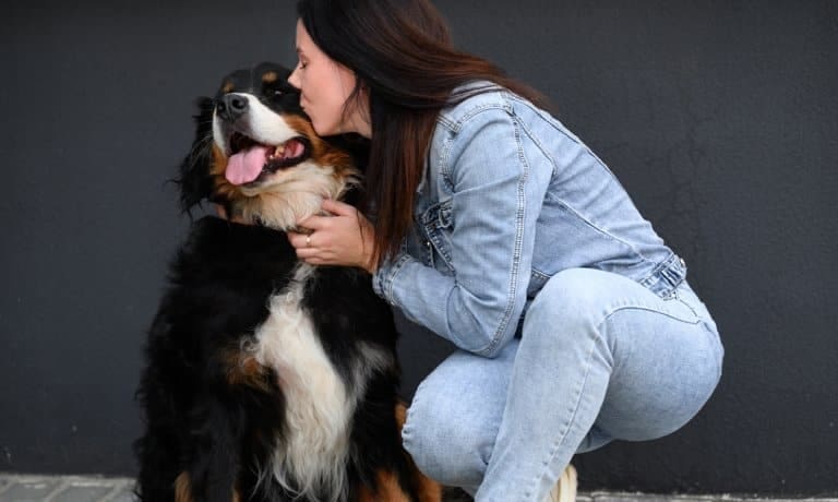 Woman in denim outfit kneeling and kissing a large black and brown dog with tongue out, sitting by a dark wall.