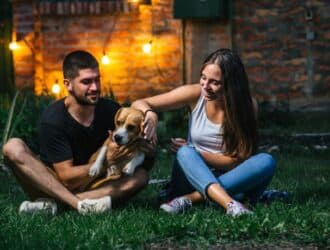 A couple plays with their dog in their backyard at night. String lights hang along the brick wall behind them.