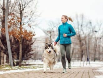 A woman wearing a light blue coat and gray leggings is jogging beside a large, fluffy dog. Snow lightly covers the ground.