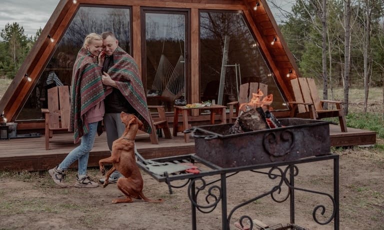 A couple wrapped in a blanket stands outside a tiny home with a brown dog. They have a fire going on a camp stove.