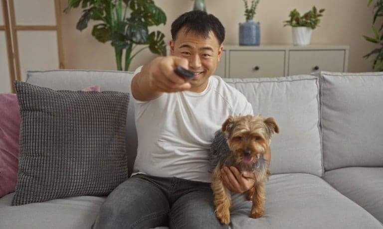 A man smiles as he holds his pet dog in one hand and a remote in the other. He leans over to turn on a television.