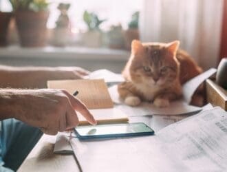 A man doing paperwork and using his phone while his cat loafs next to him on the table, its paws resting on documents.