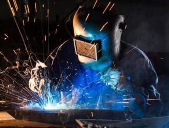 A worker performing argon welding on metal with a torch as bright sparks flare from the point of contact.