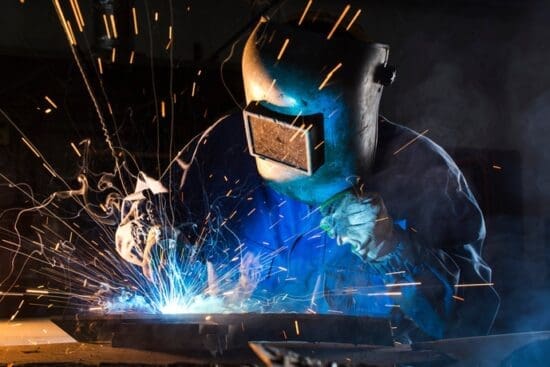 A worker performing argon welding on metal with a torch as bright sparks flare from the point of contact.