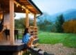 A woman sits on cabin porch steps with a small brown dog, overlooking a green field and misty mountains at sunrise.