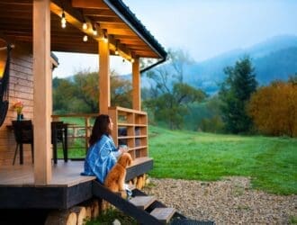 A woman sits on cabin porch steps with a small brown dog, overlooking a green field and misty mountains at sunrise.