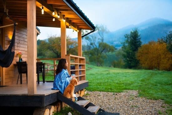 A woman sits on cabin porch steps with a small brown dog, overlooking a green field and misty mountains at sunrise.