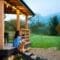 A woman sits on cabin porch steps with a small brown dog, overlooking a green field and misty mountains at sunrise.