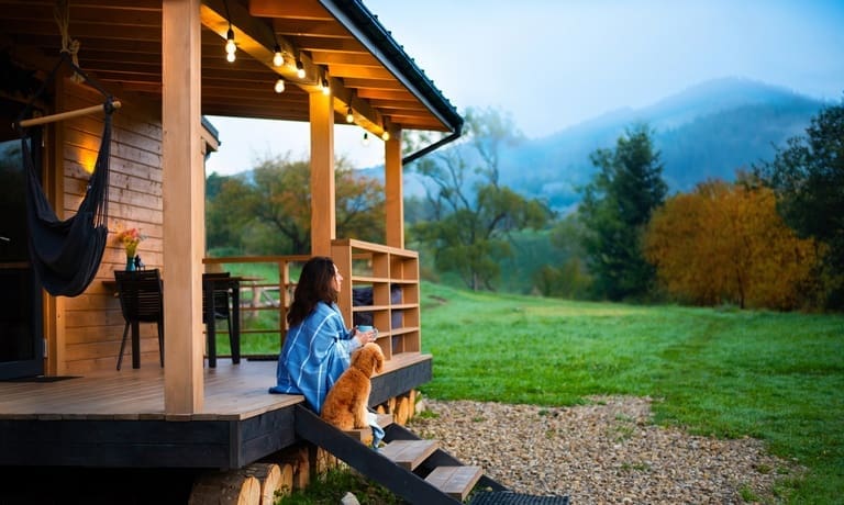 How To Plan a Cabin Getaway With Your Dog 1 A woman sits on cabin porch steps with a small brown dog, overlooking a green field and misty mountains at sunrise.