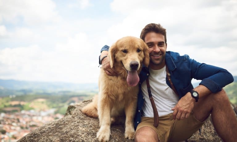 A dog owner sitting next to their panting golden retriever while both sit on a large rock overlooking a town.