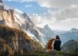A person sitting next to their dog while resting in front of a scenic mountain view on an outdoor trail.