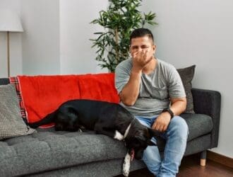 A man sits on a gray couch next to a black dog. He is pinching his nose and making a disgusted face.