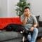 A man sits on a gray couch next to a black dog. He is pinching his nose and making a disgusted face.