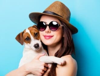 A woman wearing sunglasses and a straw hat holding a small brown and white dog against a bright blue background.