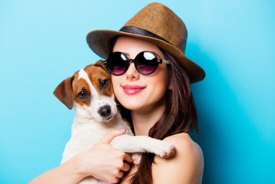 A woman wearing sunglasses and a straw hat holding a small brown and white dog against a bright blue background.