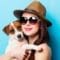 A woman wearing sunglasses and a straw hat holding a small brown and white dog against a bright blue background.