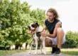 A man crouching on a park path beside a leashed dog wearing a harness, with green trees in the background.