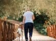 An older woman walks a medium dog across a wooden bridge surrounded by trees and greenery on a sunny day.